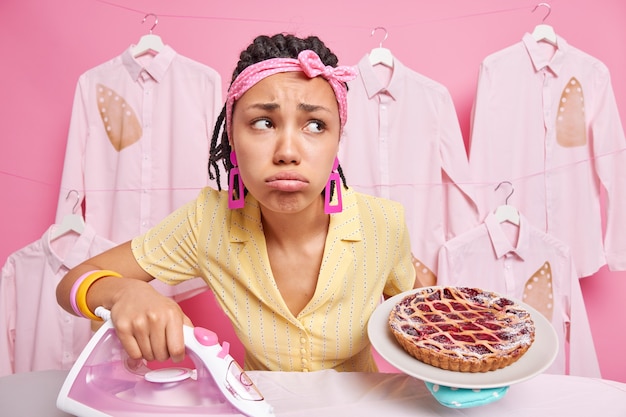 ドレッドヘアを持つ悲しい苦しめられた暗い肌の女性は、プレートに焼きたてのパイを保持します服をなでるために電気アイロンを使用します疲労感は頭にヘッドバンドを着用します