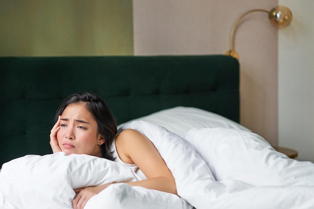 Sad and distressed young korean woman lying in her bed with pillow and warm duvet feels uneasy frowning and grimacing