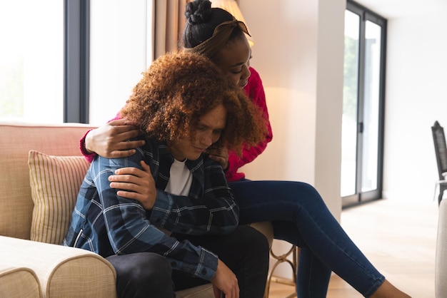 Photo sad diverse couple sitting in living room at home, embracing and comforting. depression, mental health, relationship, support, togetherness, lifestyle and domestic life, unaltered.