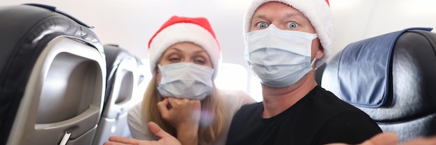 Sad and drunk man and woman in protective medical masks and santa claus red caps flying in airplane