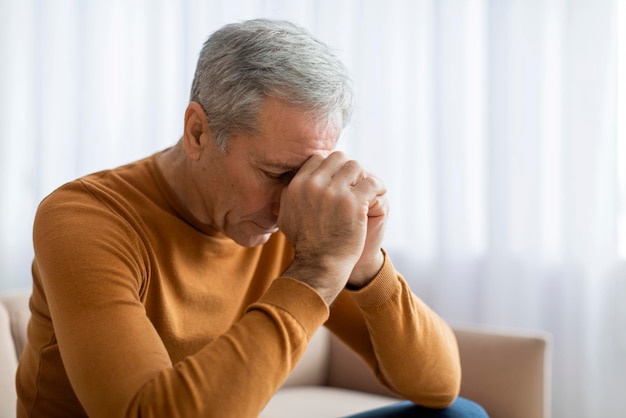 Sad elderly man sitting on sofa at home touching head