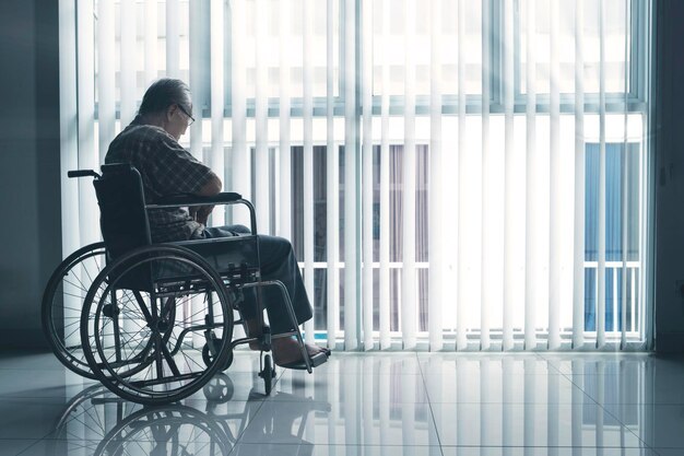 sad elderly man sitting in the wheelchair while looking out the window