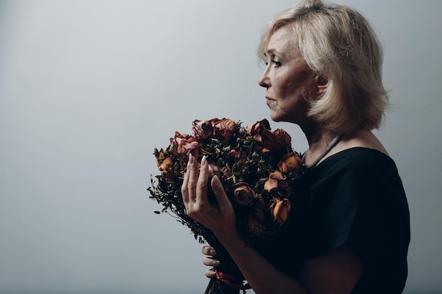 Sad elderly woman old granny touching bouquet of withered rose flowers. Gray haired mature lady in depression. Concept of old aging. Studio shoot isolated on gray background. Copy space