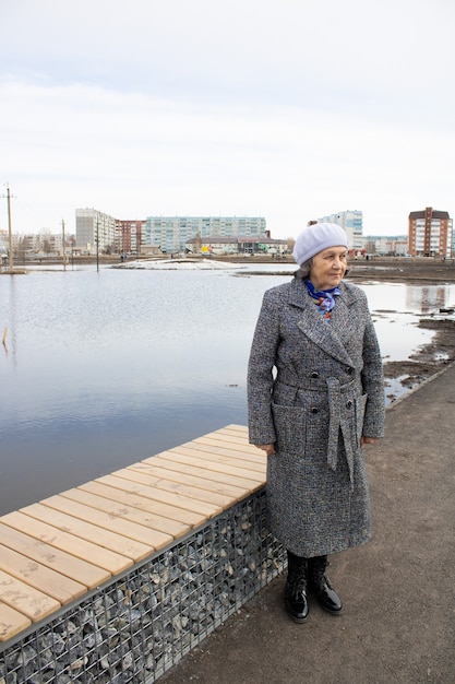 A sad elderly woman in warm clothes stands by a bench in the fresh air by the water Flooding