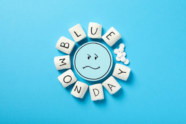 Sad emoji pills and marshmallows with text Blue Monday on blue background top view