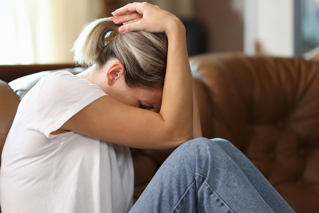 sad exhausted woman holding her head with her hands while suffering on couch at home