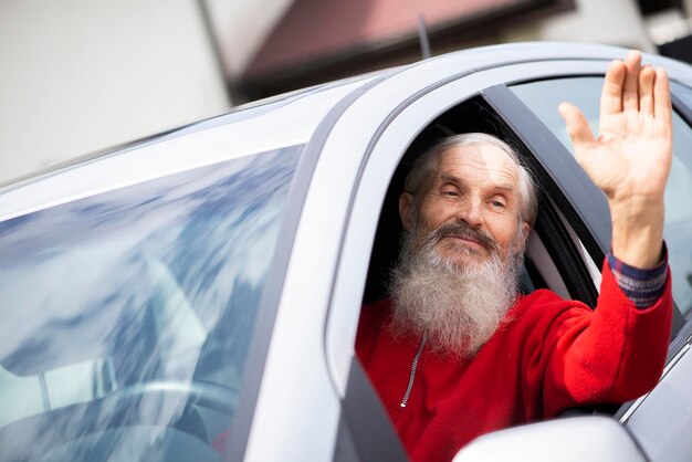 Foto testa triste uomo anziano barbuto che agita la mano attraverso la finestra di un'auto grigia