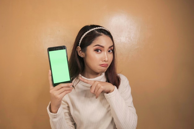 Грустное лицо молодой азиатки, указывающей на мобильный телефон в белой рубашке