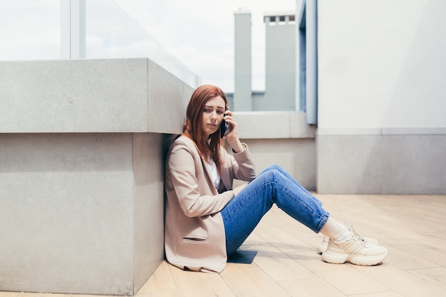 Sad female office worker sitting on the balcony floor of a modern office