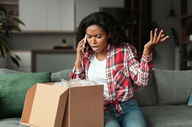 Sad frustrated millennial african american woman opening cardboard box talking on phone swearing