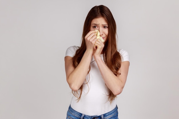 Triste donna frustrata che piange e nasconde il viso tra le mani sconvolta per la perdita di lutto sentendo un'emozione disperata che indossa una maglietta bianca indoor studio girato isolato su sfondo grigio