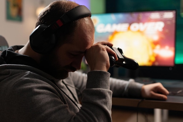 Sad gamer losing video games after playing with controller and headphones on computer. Man feeling disappointed about lost game, using controller and headset. Player gaming with equipment