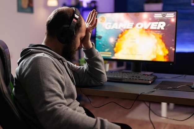 Sad gamer losing video games play on computer. Man playing online games with headphones and controller, feeling disappointed about lost game. Player using joystick and monitor.