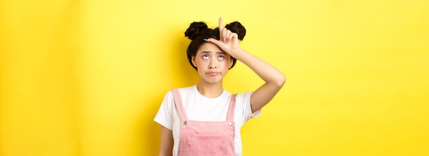 Sad girl showing loser sign on forehead and sulking upset feeling disappointed in herself standing o