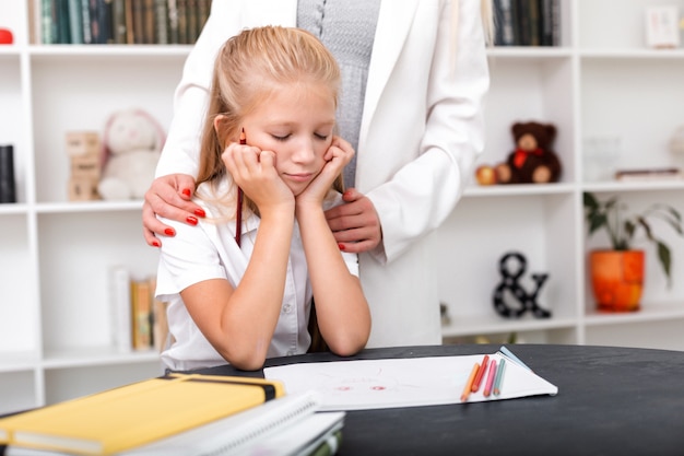 Sad girl sits at the table, draws, her mom hugs her shoulders and comforts