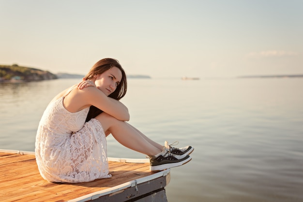 Sad girl sitting on the pier hugging her knees looking into the distance