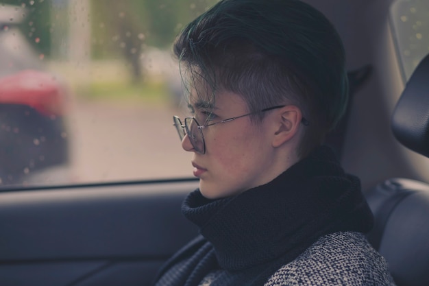 Sad girl with glasses sits in a car in autumn on a rainy day.