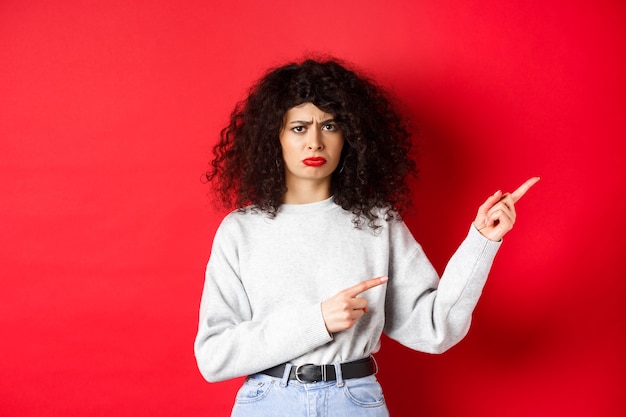 Sad and gloomy brunette woman with curly hair, frowning and looking disappointed, pointing fingers right at logo, standing on red background.