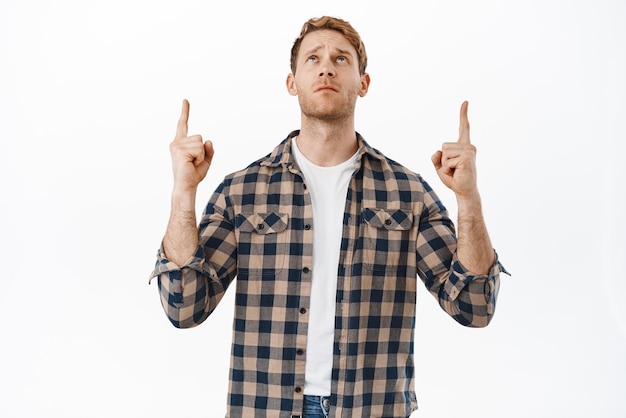 Sad and gloomy redhead man looking with regret at something unfair frowning coy pointing and staring up at promotional offer text on copyspace standing over white background