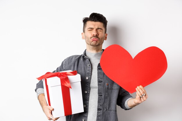 Sad and heartbroken man being rejected, crying and holding red heart with gift box, breakup on Valentines day, white. Love and relationship concept.