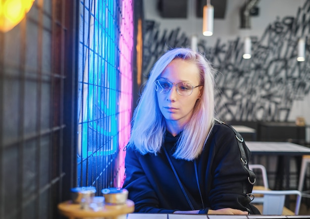 Sad hipster blonde girl in glasses sitting in bar near the neon 