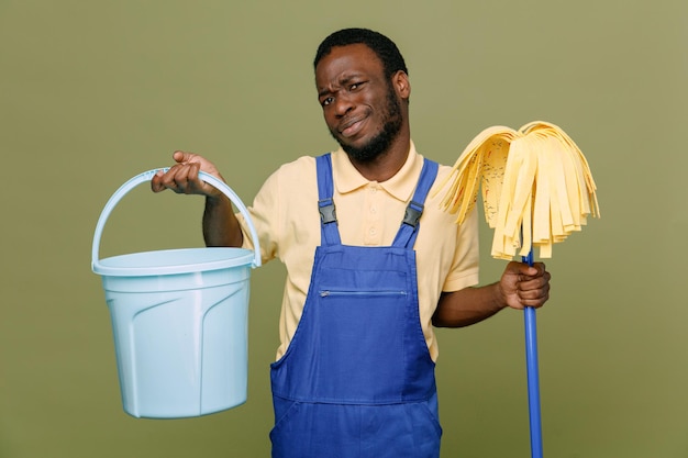 sad holding bucket with mop young africanamerican cleaner male in uniform with gloves isolated on green background