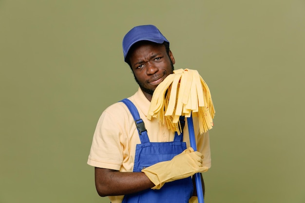 Triste holding mop giovane pulitore afroamericano maschio in uniforme con guanti isolati su sfondo verde