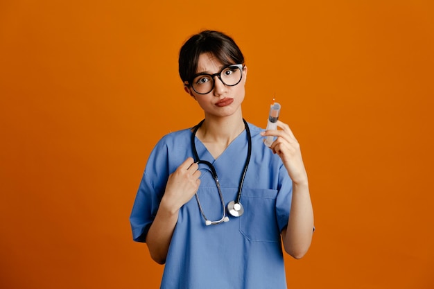 Sad holding syringe young female doctor wearing uniform fith stethoscope isolated on orange background