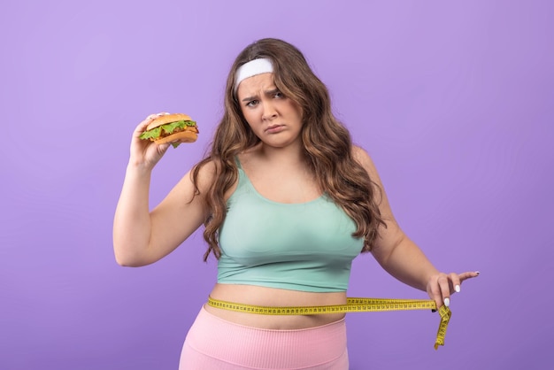 ハンバーガーを手にしたスポーツウェアの悲しい空腹の若いヨーロッパのプラスサイズの女性は、彼女の腰を測定します