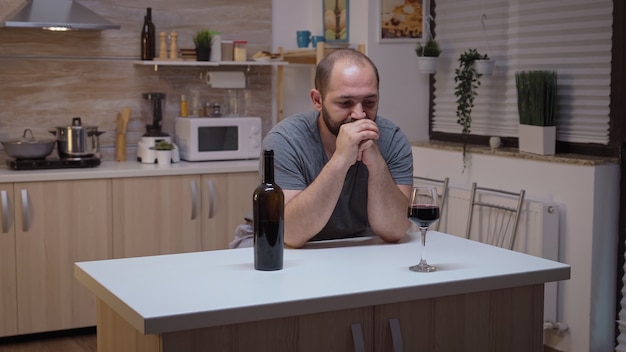 Sad husband drinking red wine sitting in the kitchen. Unhappy person suffering of migraine, depression, disease and anxiety feeling exhausted with dizziness symptoms having alcoholism problems.