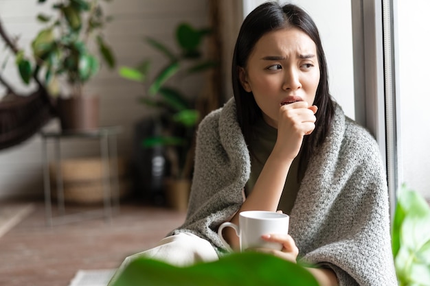 Sad ill asian girl staying on self quarantine during covid catching flu and sitting at home with cup...