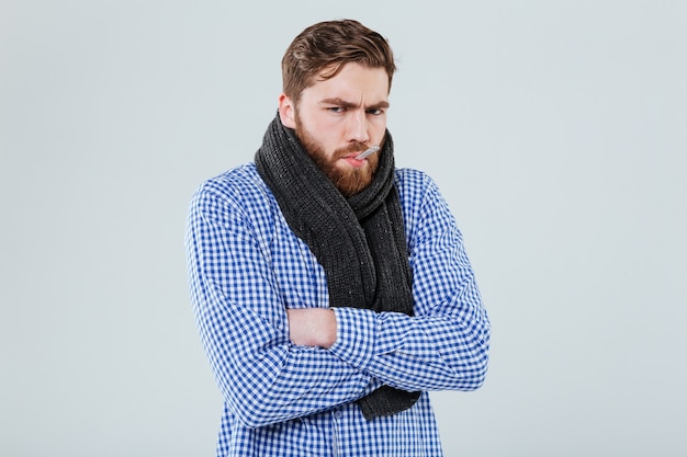 Sad ill bearded man in scarf with thermometer in his mouth over white wall