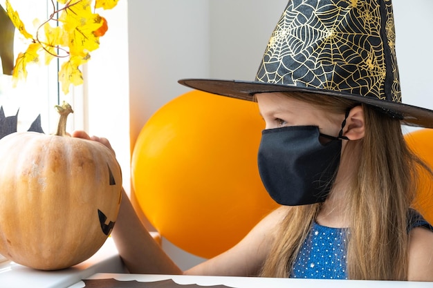 Sad kid girl in black face mask and witch costume looking at the pumpkin in decorated room