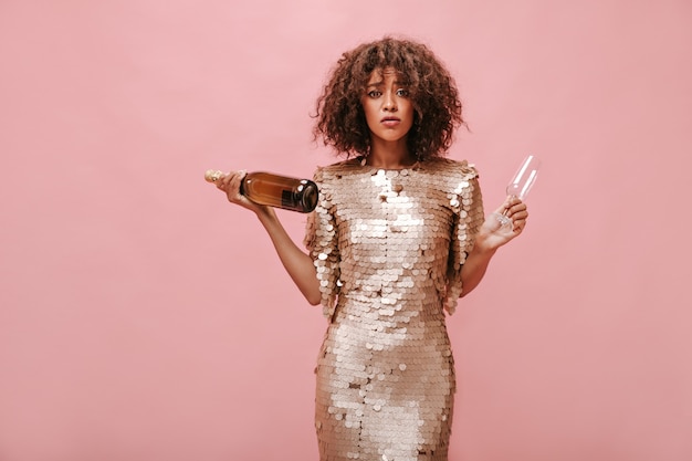 Photo sad lady in stylish beige shiny dress looking into camera and holding bottle with drink and glass on pink isolated wall..