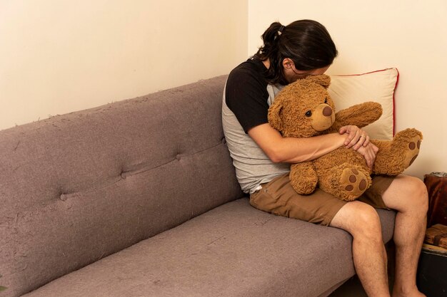 Sad Latin American man talking to a yellow teddy bear on the sofa at home	and crying