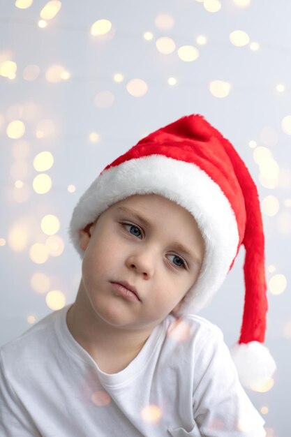 A sad little boy in a Christmas red cap on a light festive background with bokeh. Concept holidays and emotions.