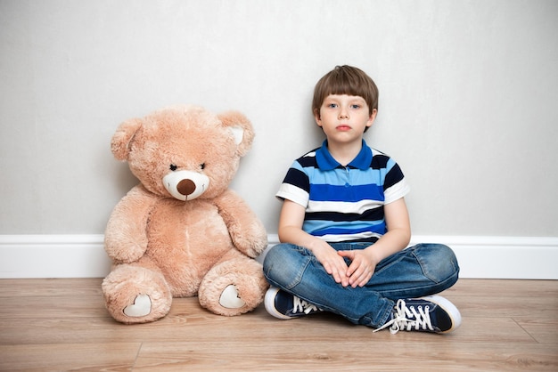 写真 大きなテディベアを持った悲しい男の子が家に座っています。子供の孤独と悲しみ。捨てられた不必要な子供たちの問題