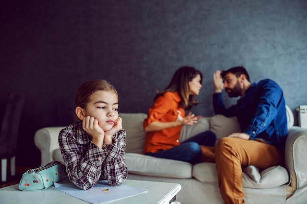 Photo sad little girl leaning on the desk and listening to parents arguing and yelling