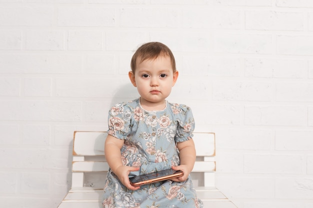 슬픈 어린 소녀는 드레스에 나무 벤치에 앉아 그녀의 손에 스마트 폰을 보유하고 있습니다.
