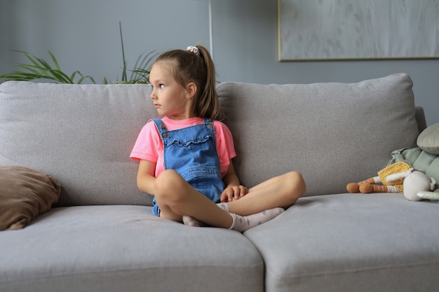 Sad little girl sitting on sofa alone at home.