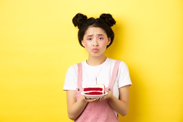 Sad and lonely birthday girl frowning upset, holding birthday cake with candle, making wish, standing on yellow background