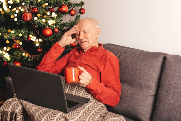 Photo sad lonely elderly caucasian man  with laptop sitting on a sofa near a christmas tree with tea mug chatting with relatives online. self-isolation, holiday mood.