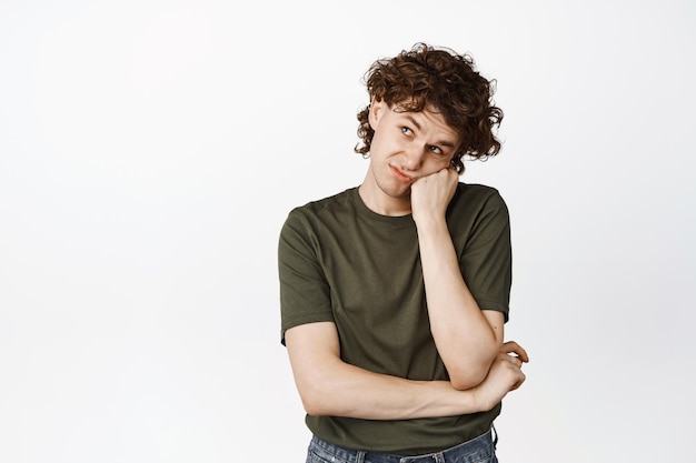 Sad and lonely young curly guy looking up lean head on hand and staring aside bored standing over white background