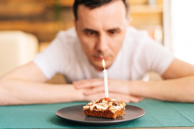 Sad lonely young man in festive hat celebrating birthday alone, sitting at the birthday cake and looking with sad eyes on it. Concept of celebrating alone.