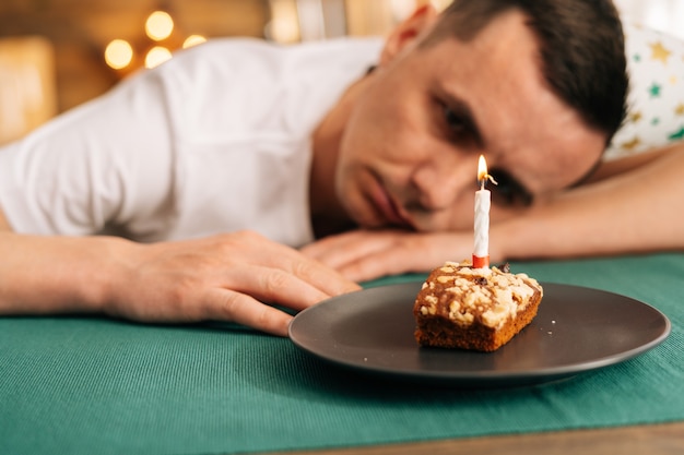 Sad lonely young man in festive hat celebrating birthday alone, sitting at the birthday cake and looking with sad eyes on it.