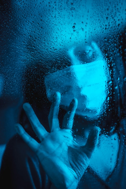 Photo sad look of a young caucasian brunette with face mask looking at a rainy night in the covid19 quarantine