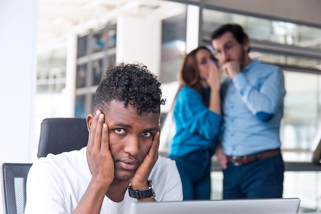 A sad looking black man is working on desk and there are two\
colleagues are gossiping at background behind of him