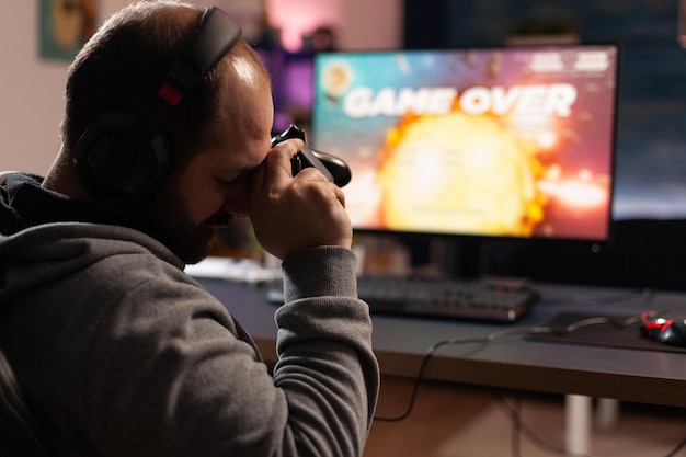Sad man losing video games after playing with joystick and headphones on computer. Gamer feeling disappointed about lost game, using controller and headset. Person gaming with equipment