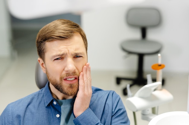 Sad man sitting in dental chair suffering from acute tooth pain rubbing his cheek close up