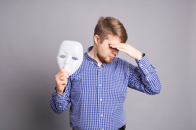 Sad man taking off plain white mask revealing face, gray wall